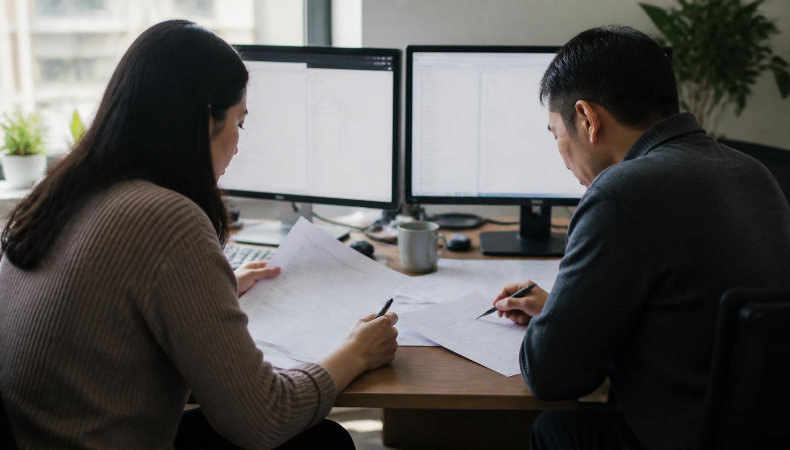 A translator and reviewer working side by side at a two-monitor desk with printed pages