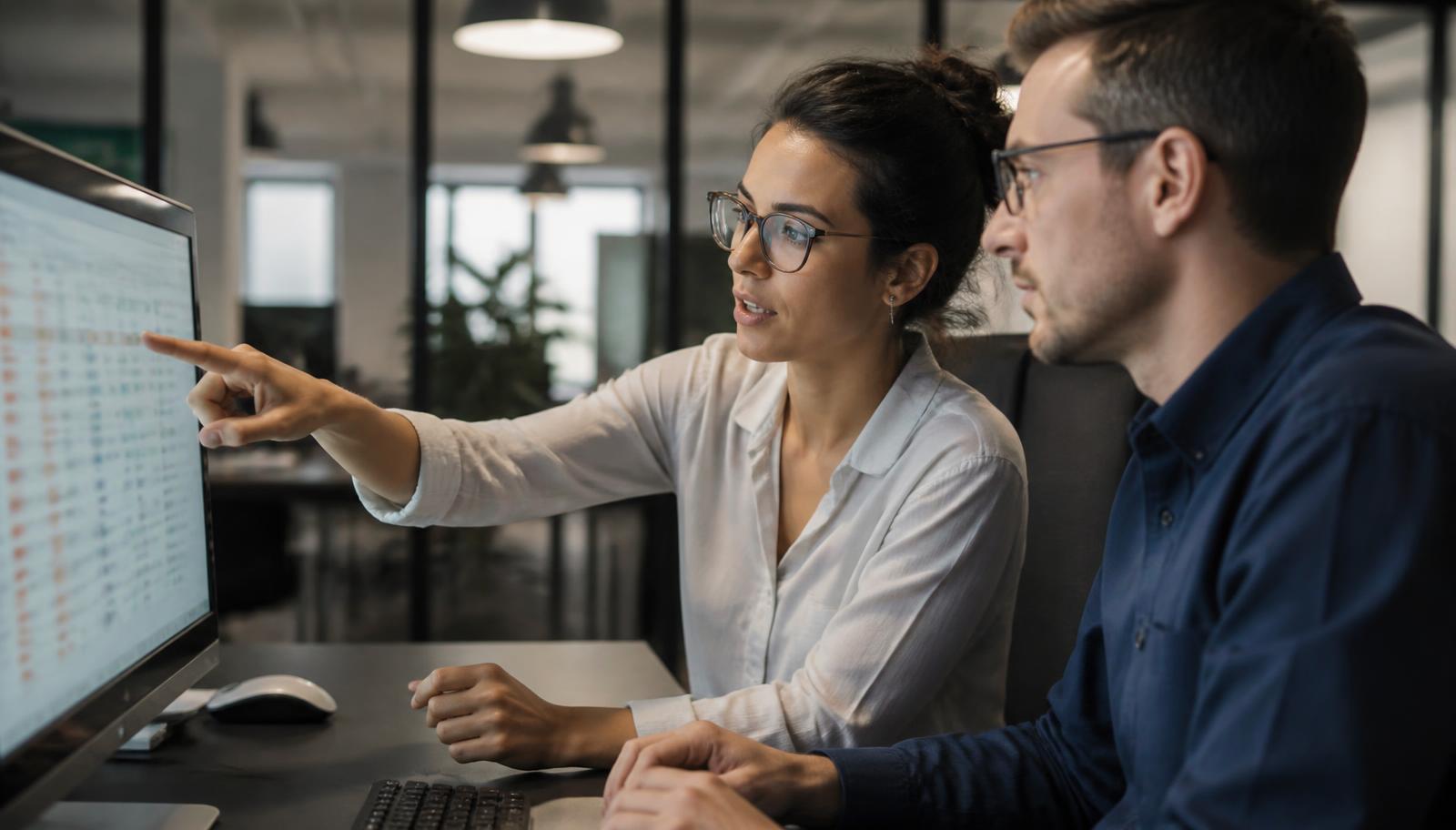 Two compliance analysts reviewing flagged items on a monitor, one pointing at a row