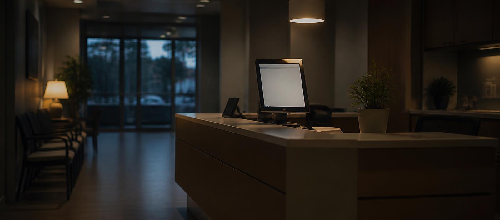 An empty clinic reception at dusk with a tidy counter and one glowing monitor