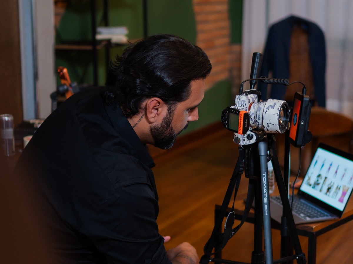 A videographer filming a talking-head clip on a tripod-mounted camera in a small studio